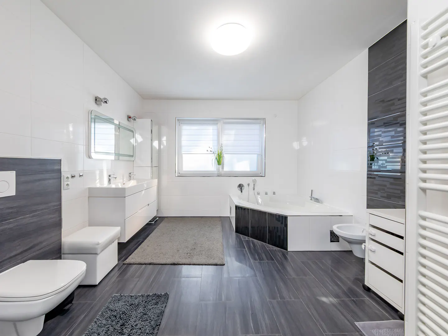 Bright bathroom with white walls, gray tile floor, and a large window. A white vanity with double sinks and a large tub are visible.