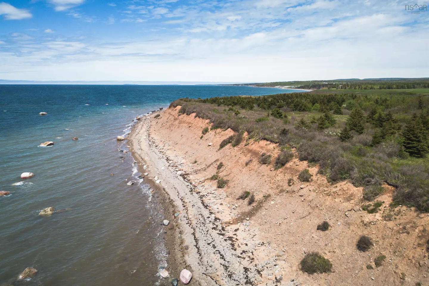 Aerial view of a rocky beach with a red clay cliff, green trees, and blue ocean under a partly cloudy sky.