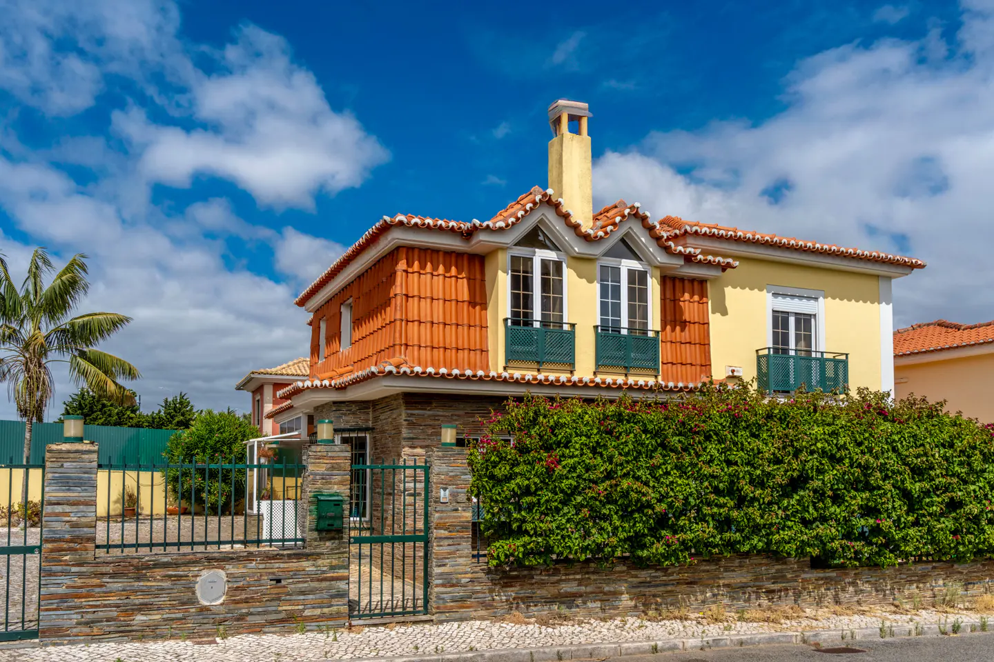 Two-story yellow house with orange tile roof, chimney, and green trim. A stone wall and gate surround the property. Blue sky with clouds.