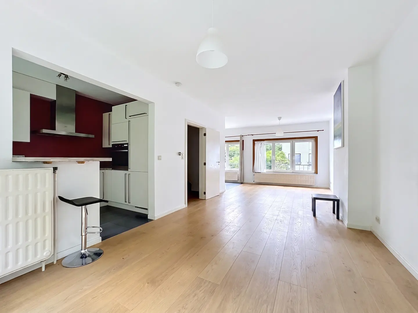 Bright, open apartment with hardwood floors, white walls, and a modern kitchen with a red accent wall. A black stool sits at the counter.