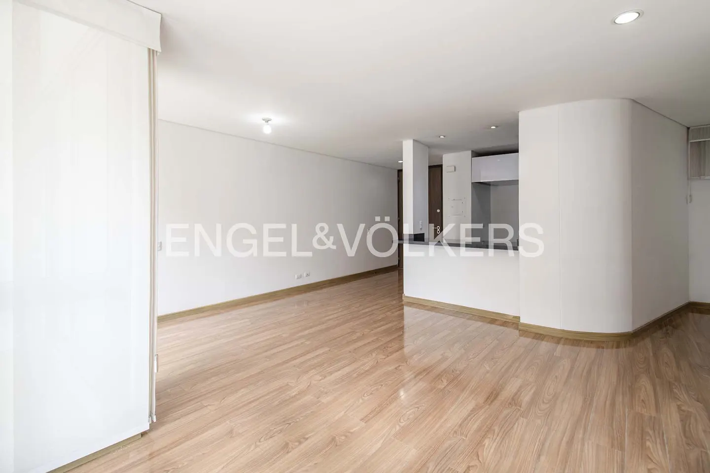 Bright, empty living room with wood floors and white walls. Kitchen area visible in the background. Engel & Völkers logo on the wall.