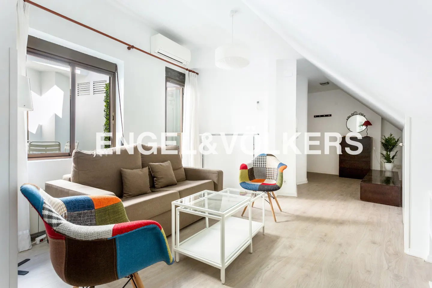 Bright living room with a beige sofa, glass coffee table, and two colorful patchwork accent chairs on light wood floors.