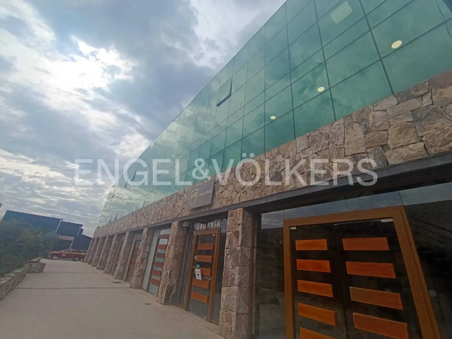 Exterior view of an Engel & Völkers office building with a glass facade and stone accents under a cloudy sky.
