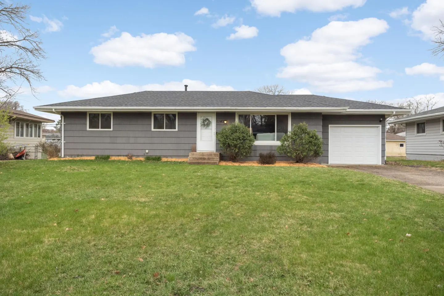 A gray ranch-style house with a white front door and garage faces a green lawn under a blue sky with white clouds.