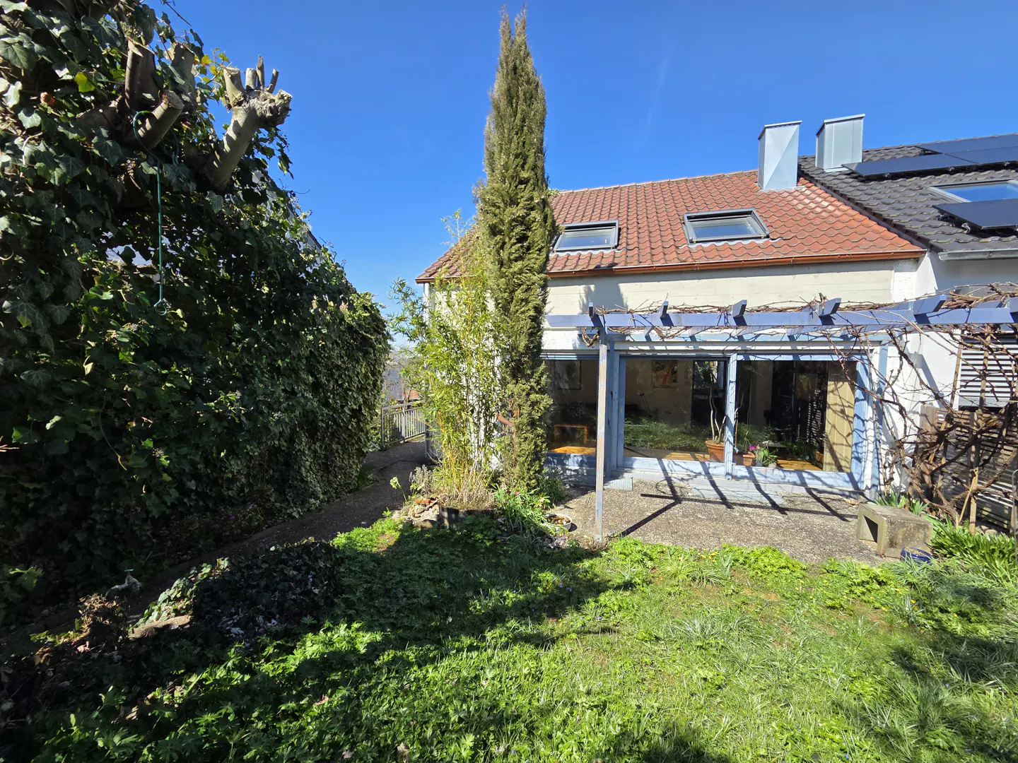 A house with a red tile roof and a green lawn on a sunny day. A tall, thin tree stands next to the house.