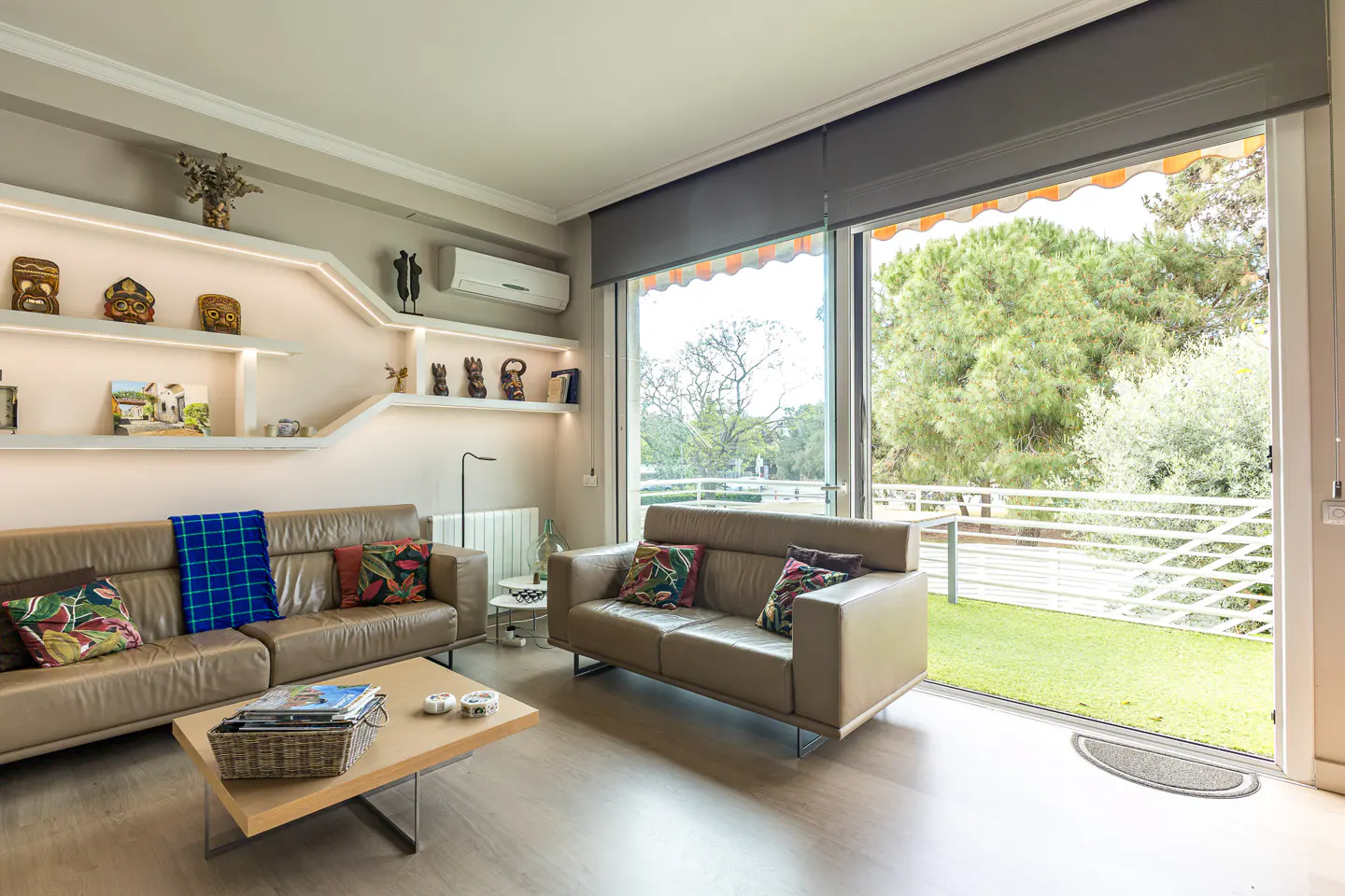 Living room with tan leather sofas, wood floor, and shelves with decorative objects. Large sliding glass doors open to a balcony with green grass.
