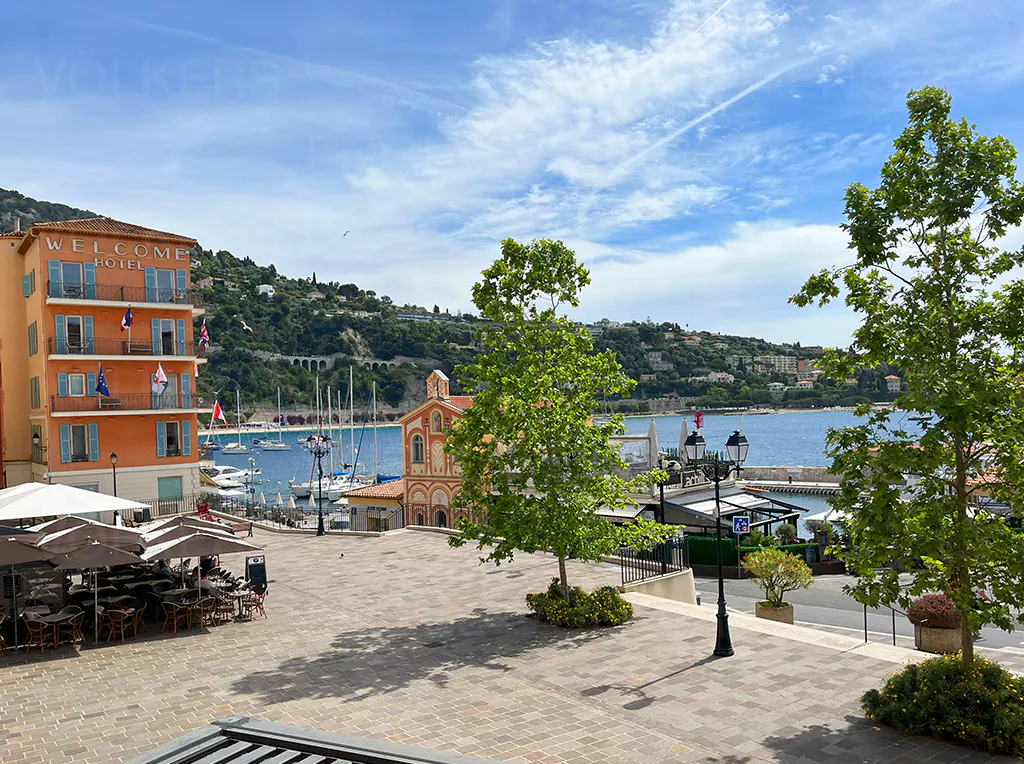 View of a sunny harbor square with the orange Welcome Hotel, outdoor cafe seating, and boats on the blue Mediterranean sea.