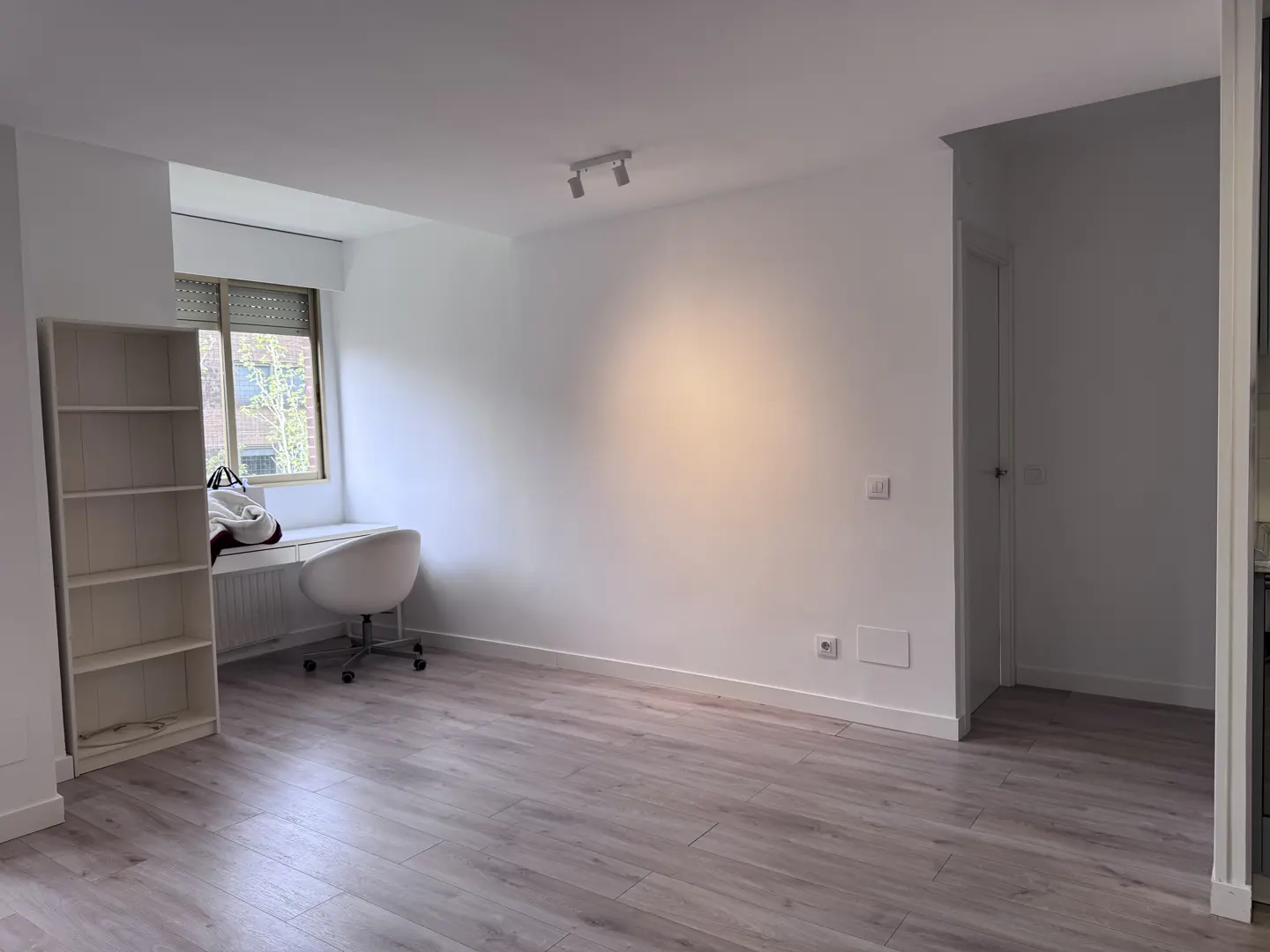 Bright, empty room with light wood floors, white walls, and a window with a desk and chair. A white bookshelf stands to the left.