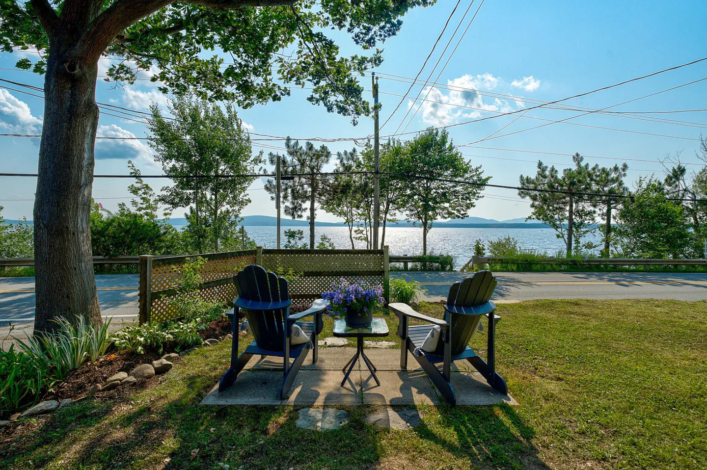 Two Adirondack chairs face a lake view, separated by a small table with flowers, on a sunny day.