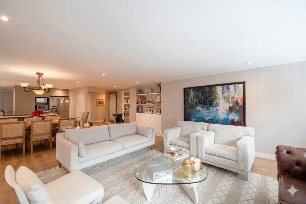 Open-concept living space with a light gray sofa, chairs, and a glass coffee table on a patterned rug. Dining area and kitchen in the background.