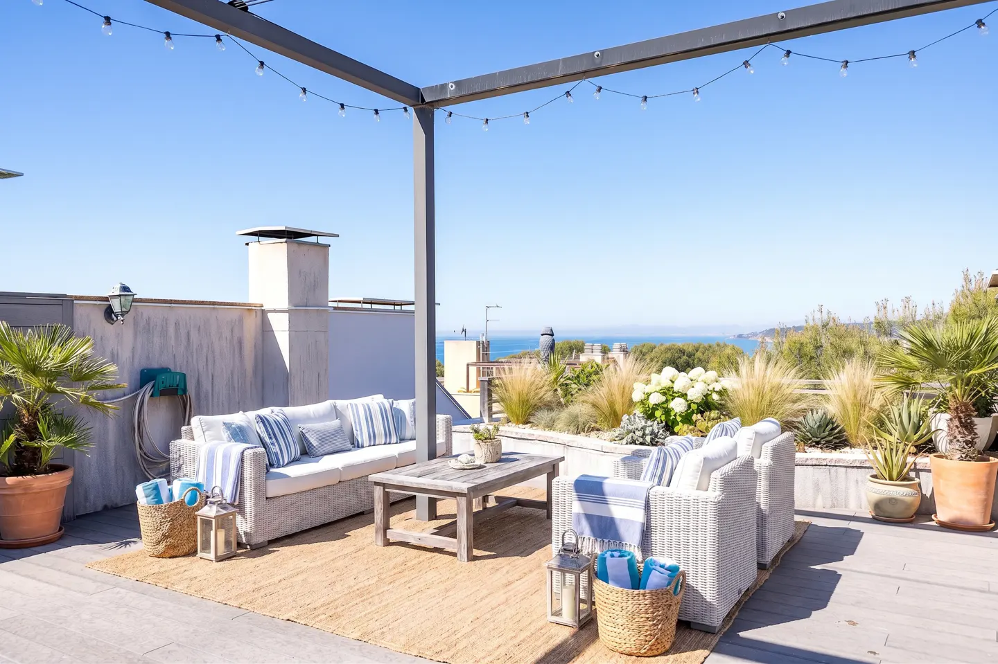 Rooftop patio with white wicker furniture, blue striped cushions, and a jute rug. String lights hang above, with ocean views in the background.