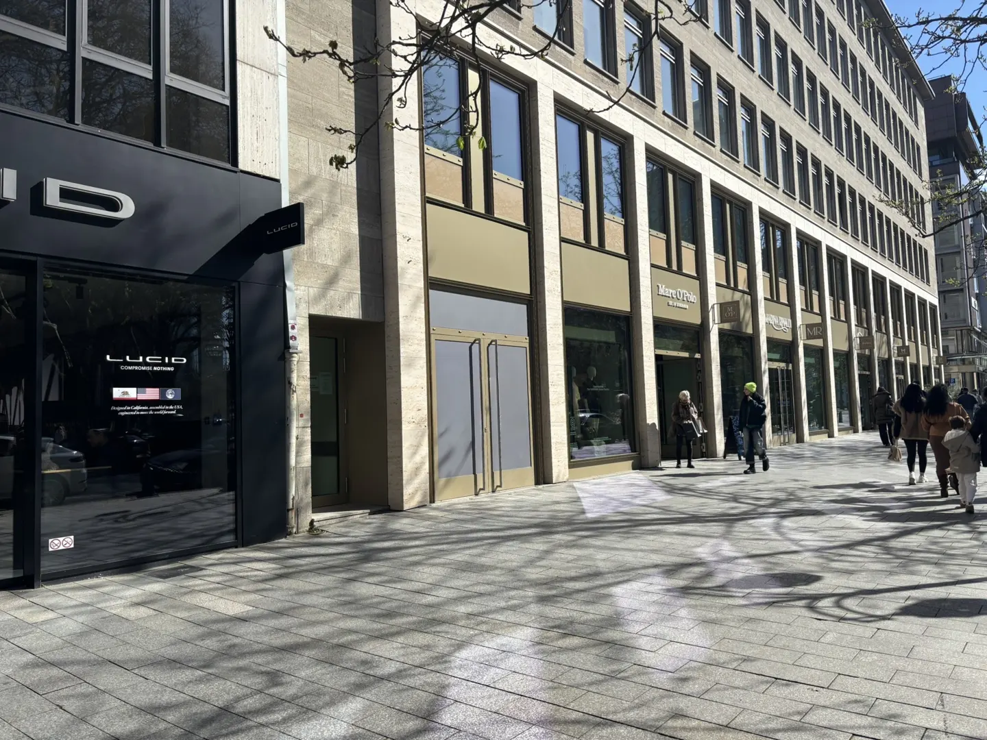 Street view of shops, including Lucid, in a tall building. People walk on the sidewalk. Tree shadows are visible on the ground.