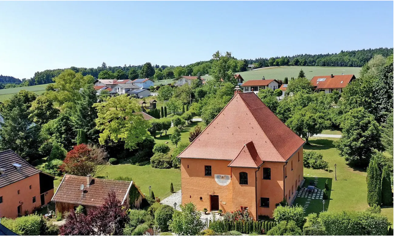 Aerial view of an orange, two-story house with a red tile roof, surrounded by green trees and lawns in a rural setting.