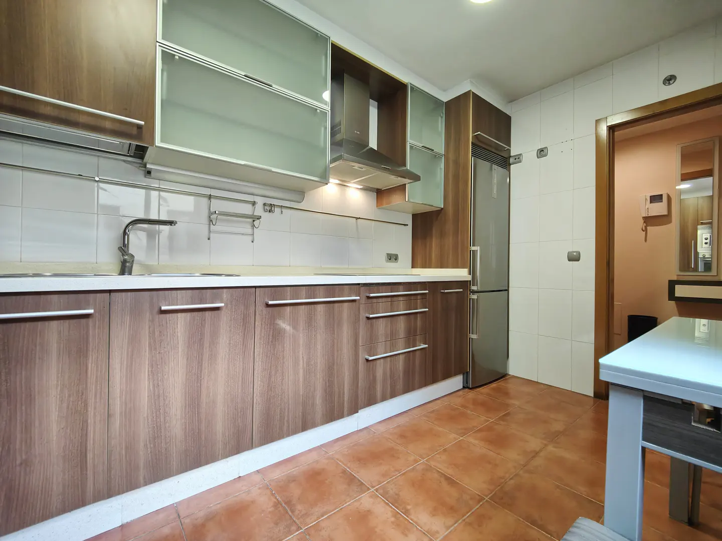 A kitchen with wood cabinets, stainless steel appliances, and terracotta tile flooring. A doorway leads to another room.
