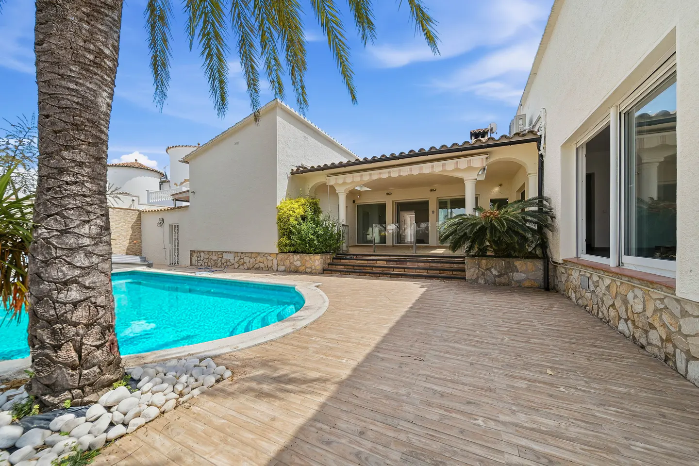 Backyard view of a white house with a blue pool, wooden deck, and palm trees under a blue sky.