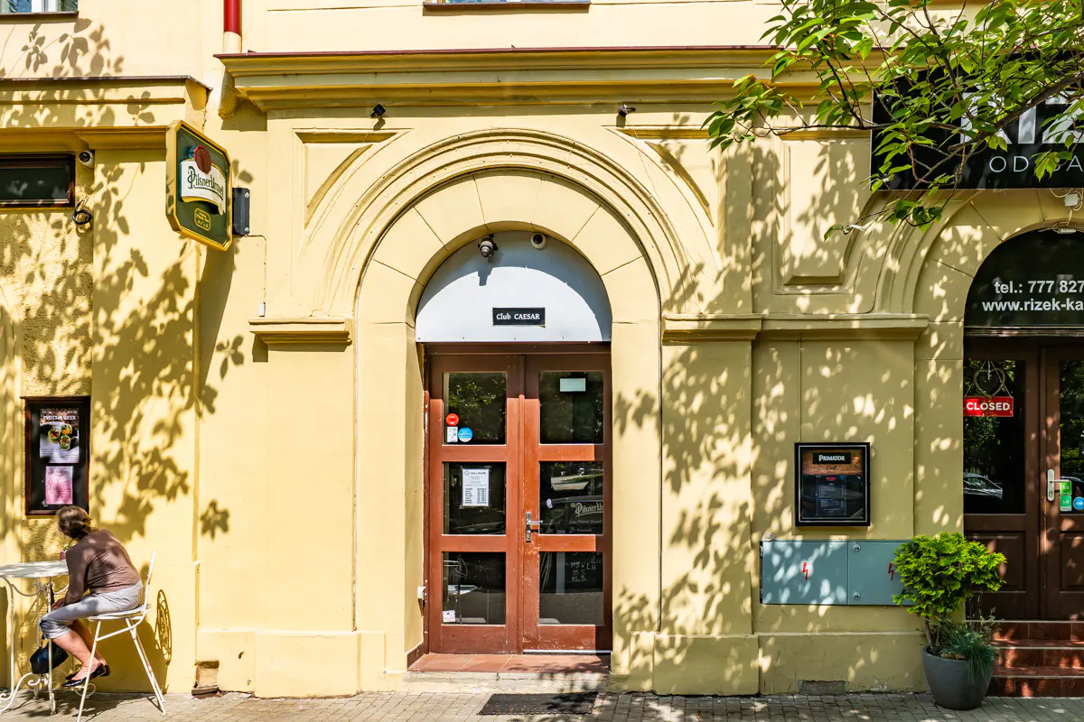 A person sits at a table outside a yellow building with a prominent arched doorway labeled Club Caesar.