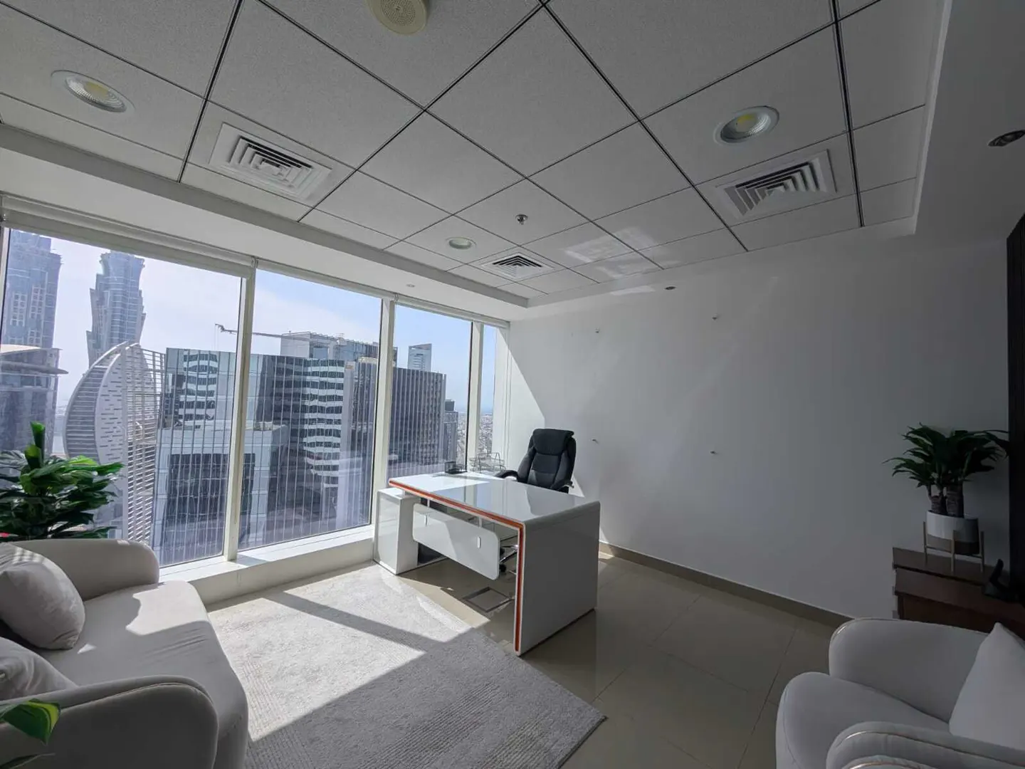 Bright office space with a white desk, black chair, and city views through large windows. A sofa and armchair provide seating.