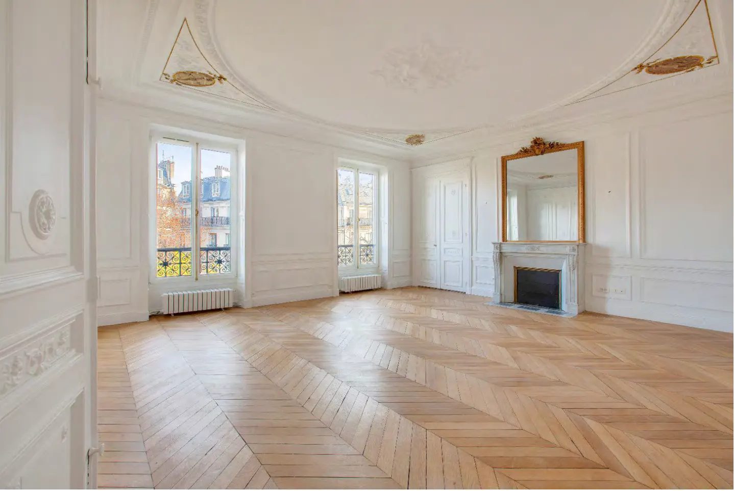 Bright, empty room with herringbone wood floors, white walls, and ornate ceiling. Windows show city view. Fireplace with gold-framed mirror.