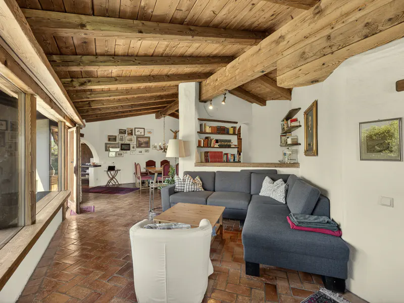 Living room with exposed wood beam ceiling, brick floors, and a gray sectional sofa. Bookshelves and framed art decorate the white walls.