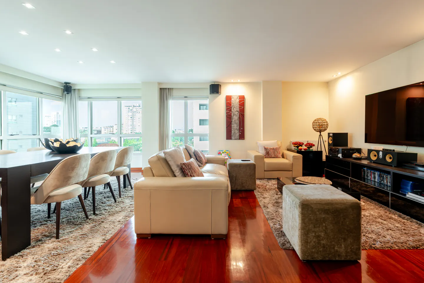 Bright, modern living room with wood floors, beige sofa and chairs, and a dining table with chairs. Large windows offer city views.