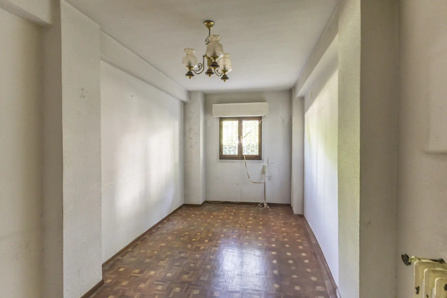 Empty room with white walls, parquet flooring, a vintage chandelier, and a window with a roller shutter.
