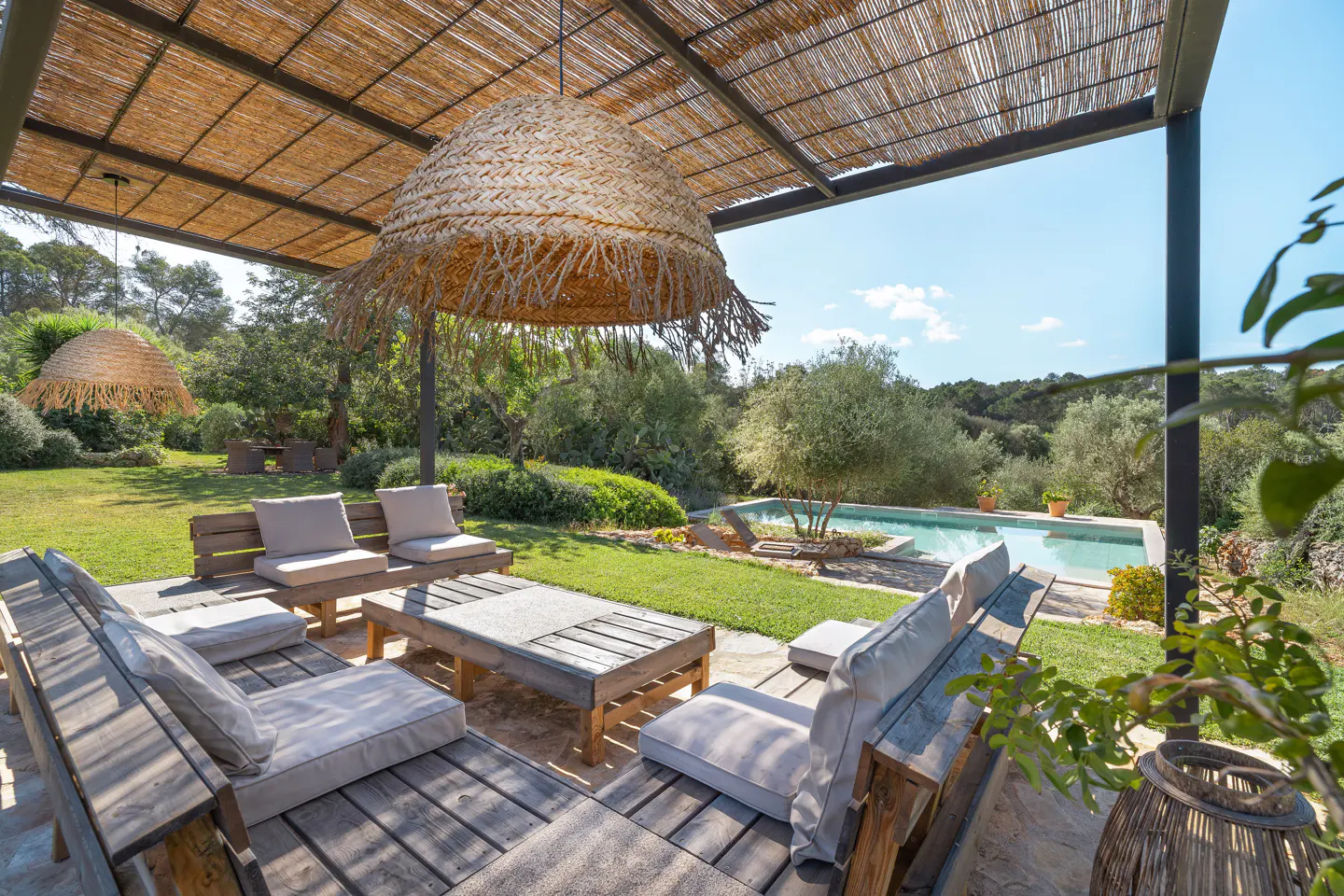 Outdoor patio with wooden furniture, straw lamps, and a pool in the background. The patio has a thatched roof and is surrounded by greenery.