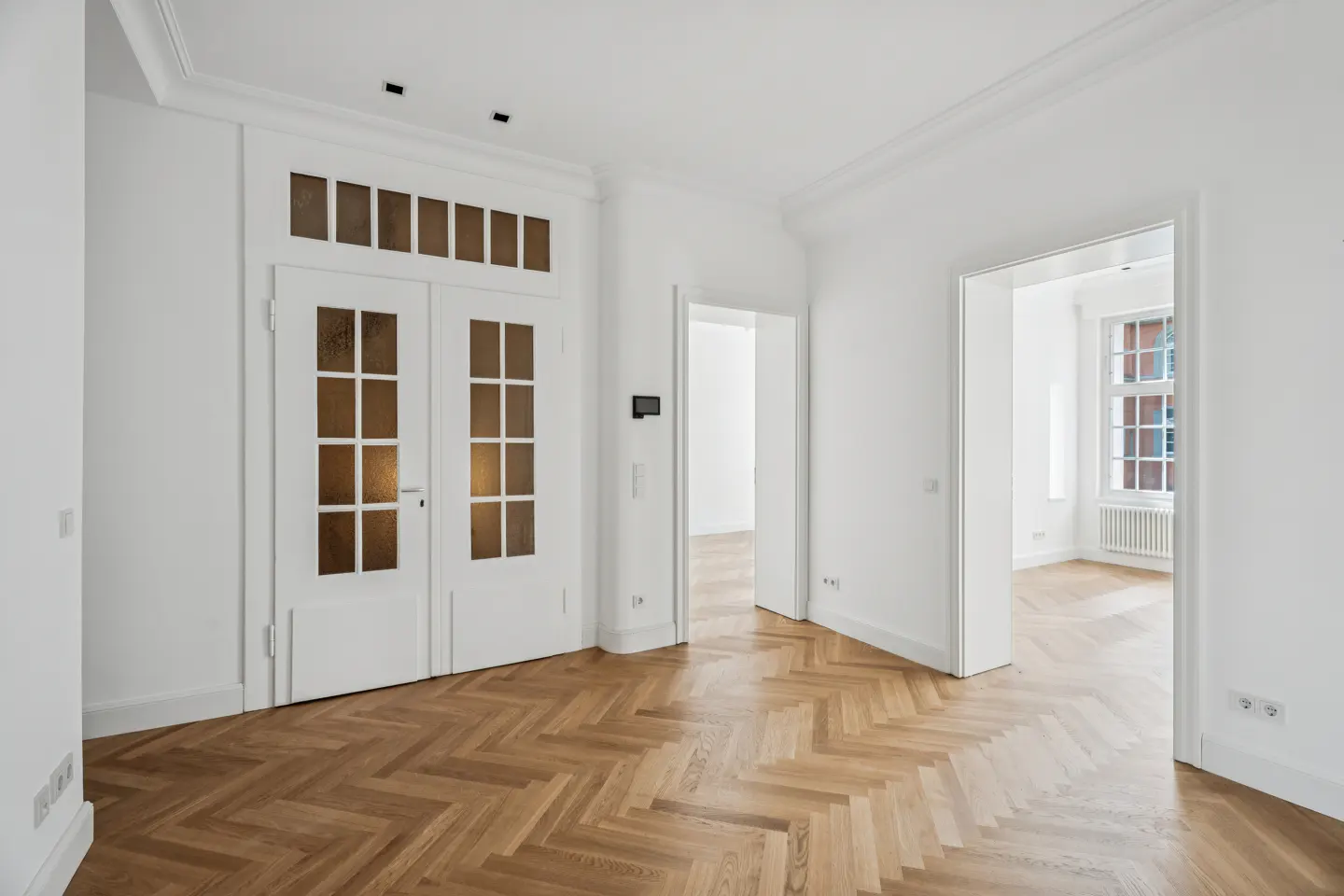 Bright, empty room with white walls, herringbone wood floors, and white double doors with glass panes leading to a hallway.