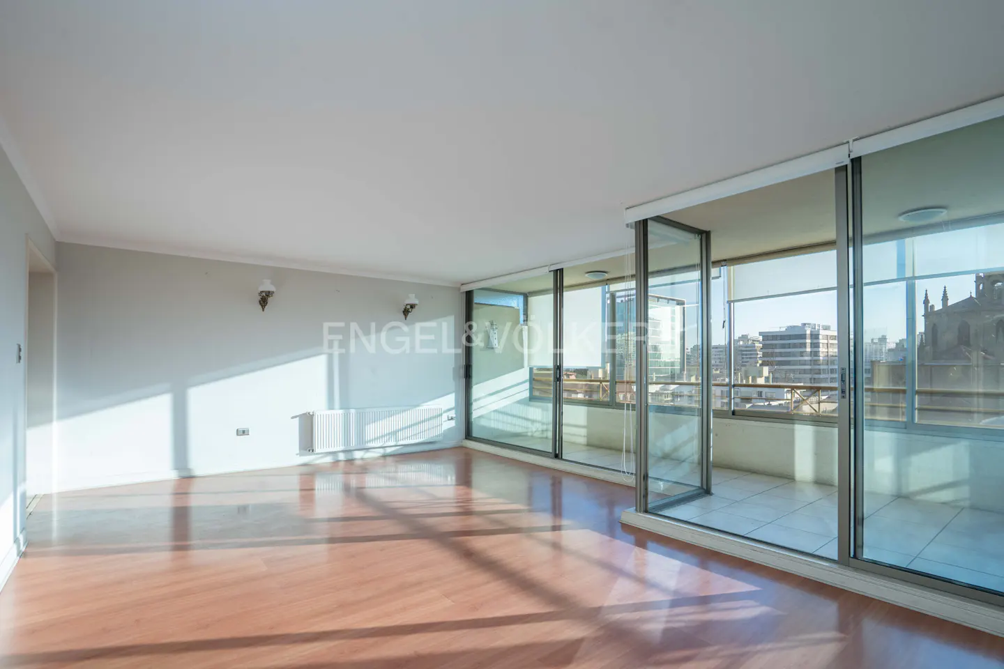 Bright, empty room with light wood floors, white walls, and large glass sliding doors opening to a city balcony view.
