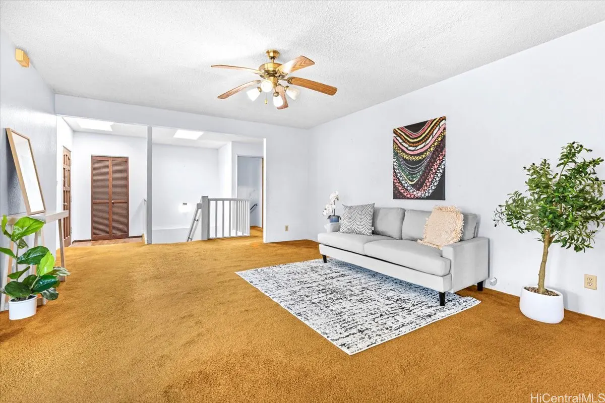 Living room with gold carpet, white walls, and a gray sofa. A ceiling fan hangs above, and a staircase leads to another level.