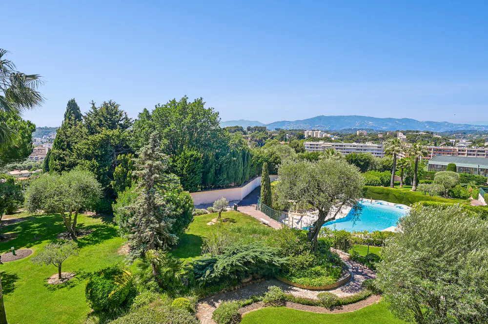 Aerial view of a lush green garden with a blue swimming pool, trees, and buildings in the background under a clear blue sky.