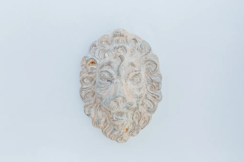 Stone lion head sculpture with a weathered finish mounted on a white wall.
