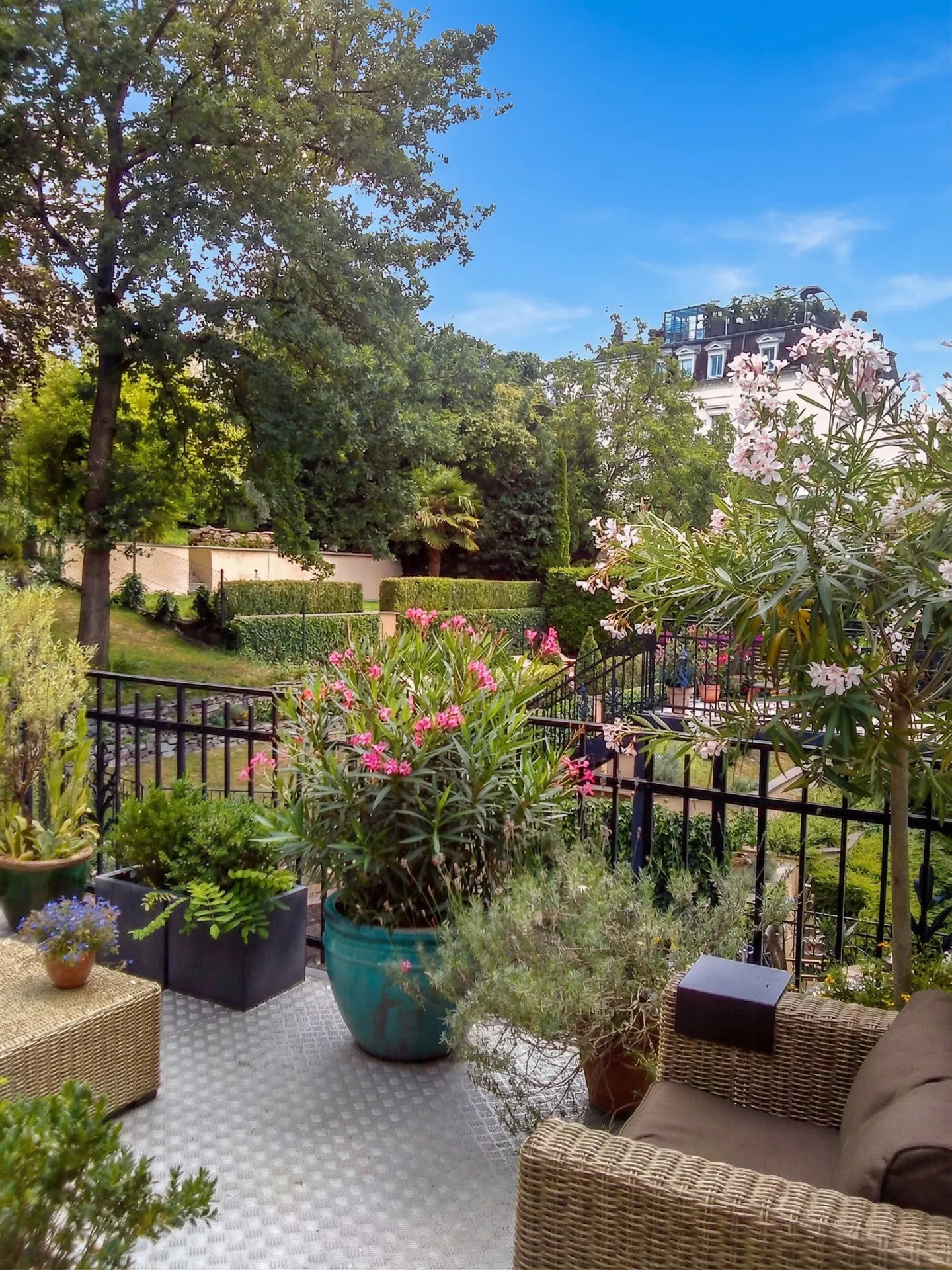 Outdoor patio with wicker furniture, potted plants, and a view of a manicured garden under a blue sky.