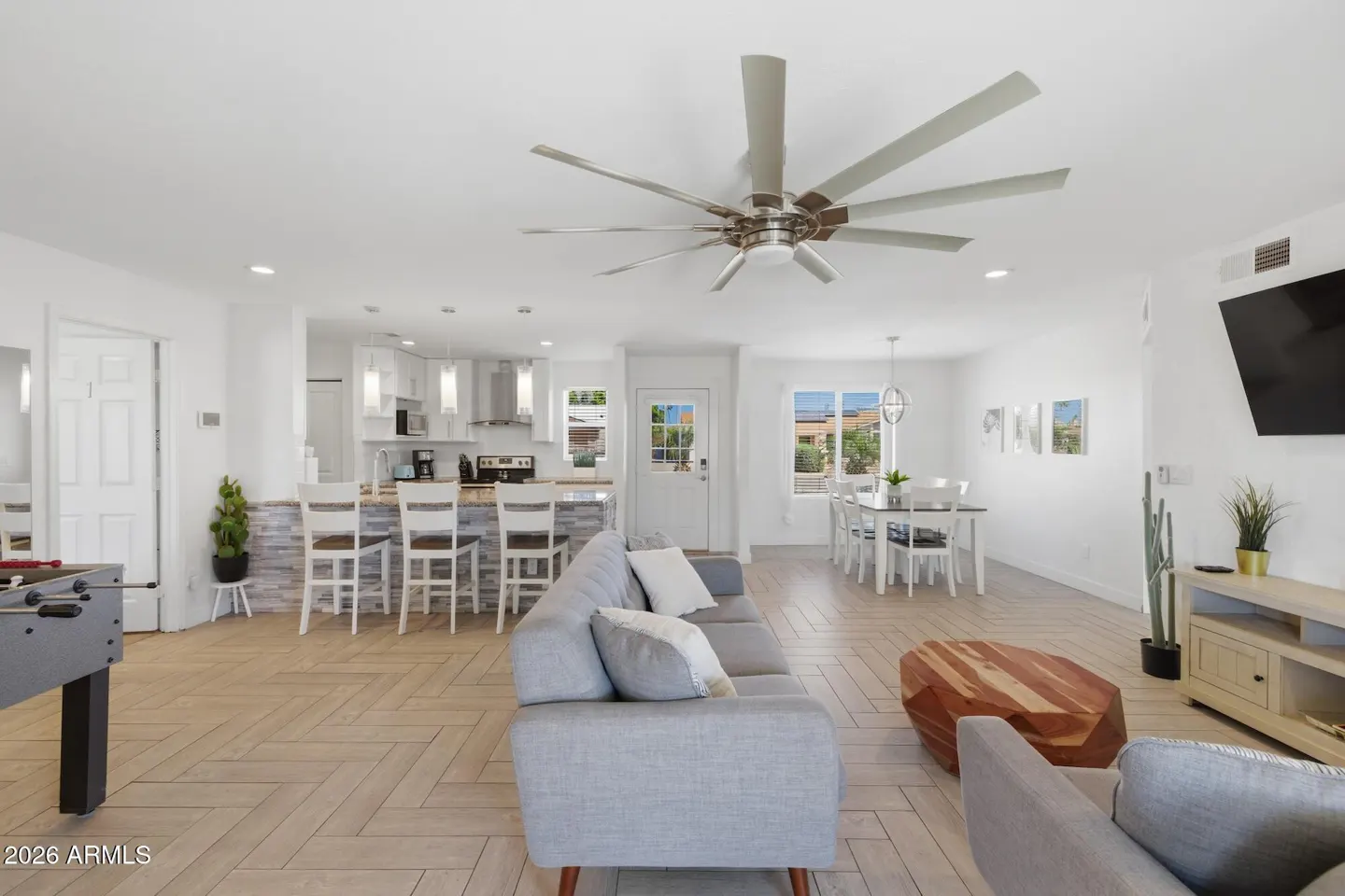 Bright, open-concept living space with a gray sofa, white kitchen, dining area, and herringbone wood floors. A large ceiling fan is overhead.