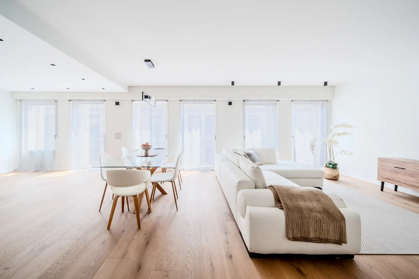 Bright, modern living room with wood floors, white walls, and large windows. A glass table with white chairs sits near a white sectional sofa.