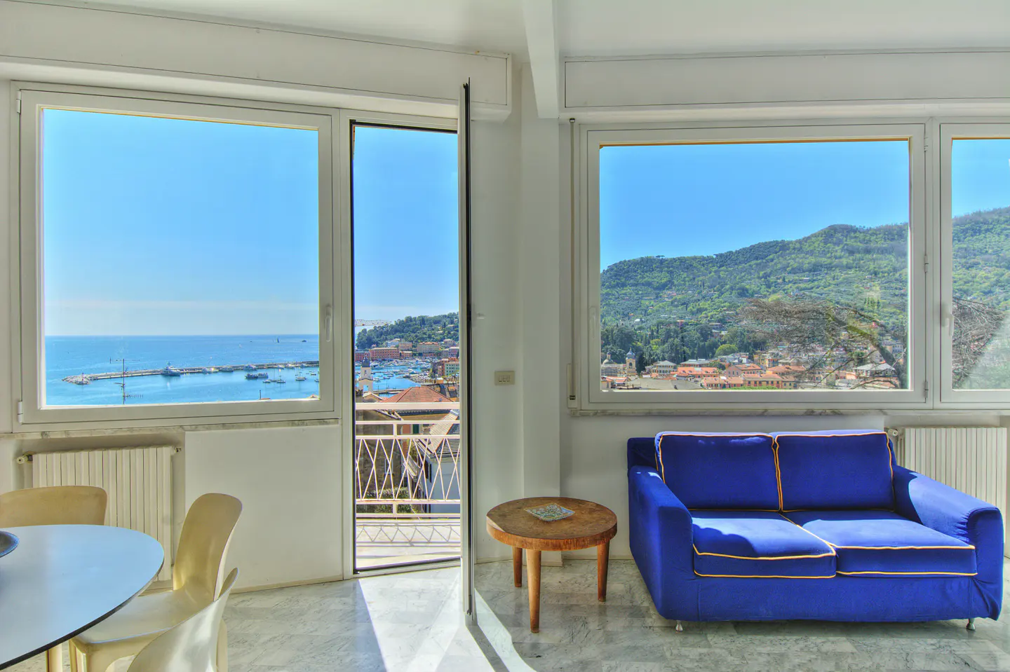 Bright apartment interior with a blue sofa, round table, and views of the sea and mountains through large windows.