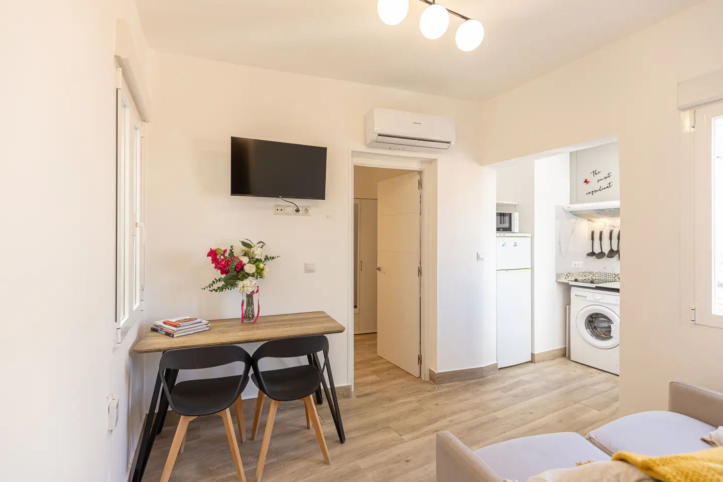 Bright studio apartment with a dining table, two black chairs, wall-mounted TV, and a view into a small kitchen.