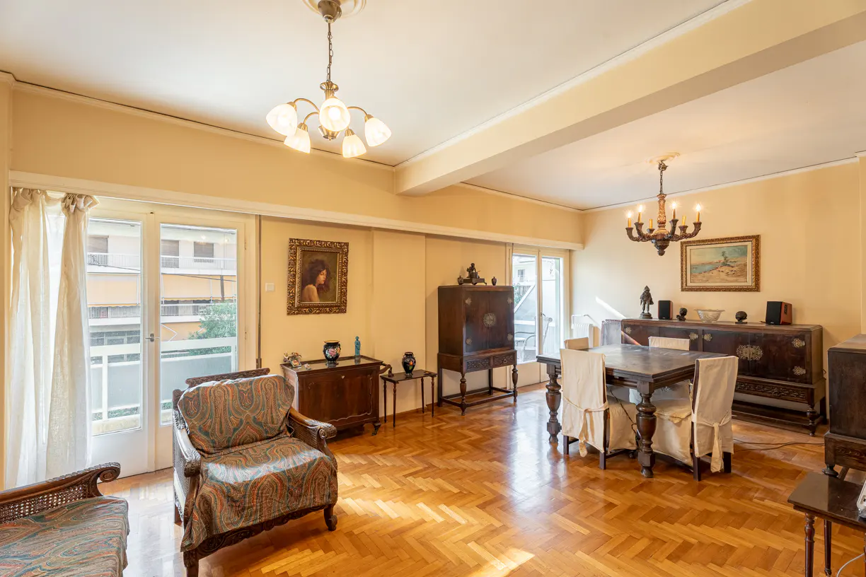 A living room with parquet floors, antique furniture, and yellow walls. Chandeliers hang from the ceiling. A balcony is visible through glass doors.