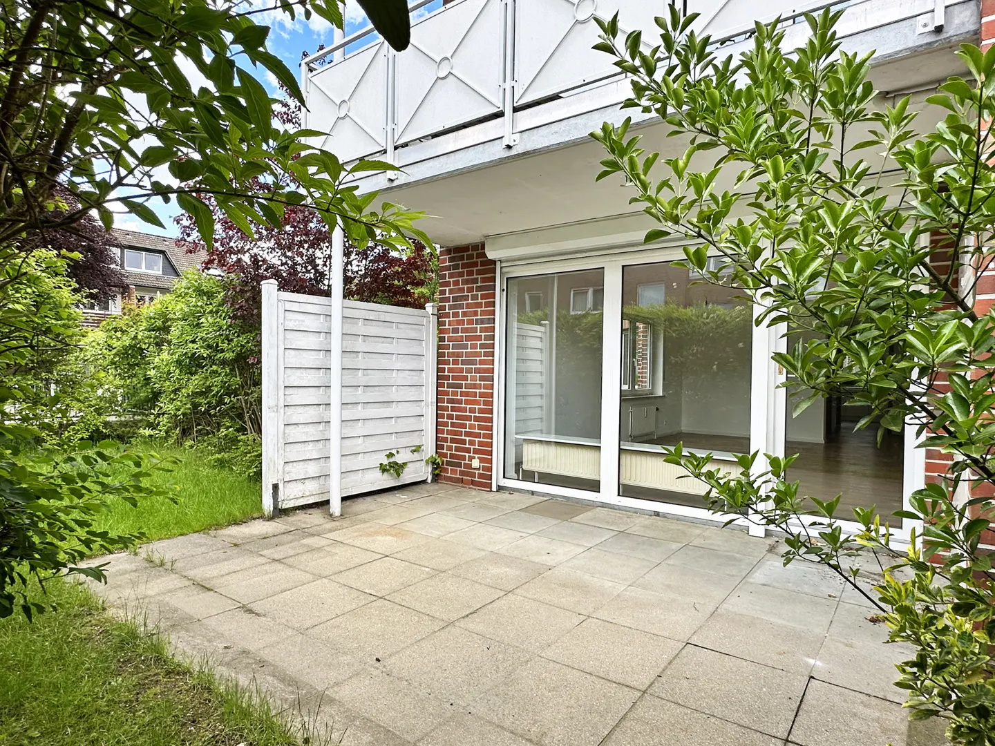 A patio with gray tiles, a white fence, and a brick wall. Green trees frame the view of a sliding glass door leading inside.