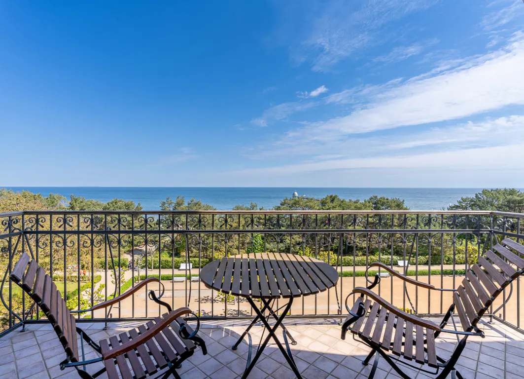 Balcony view with table and chairs overlooking the ocean and garden under a blue sky.