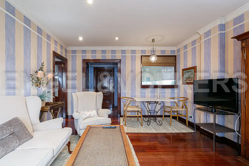 Living room with blue and beige striped wallpaper, white armchairs, a dining set, and a television on a metal stand.