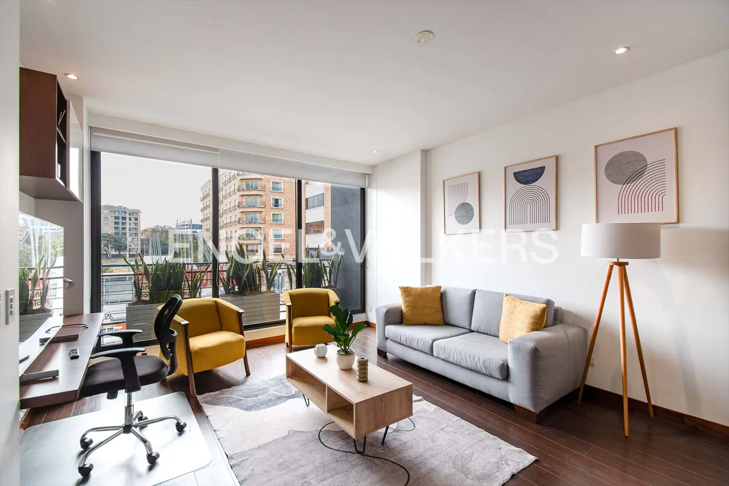 Bright living room with a gray sofa, yellow chairs, and a wood floor lamp. Large windows overlook city buildings and greenery. Modern art hangs on the white walls.