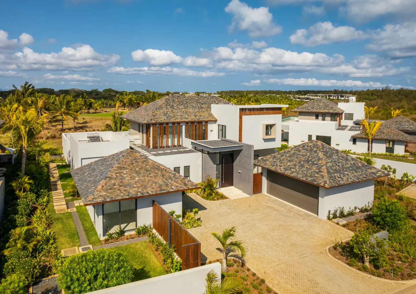 Aerial view of modern white homes with brown roofs, a driveway, and green landscaping under a blue sky with scattered clouds.
