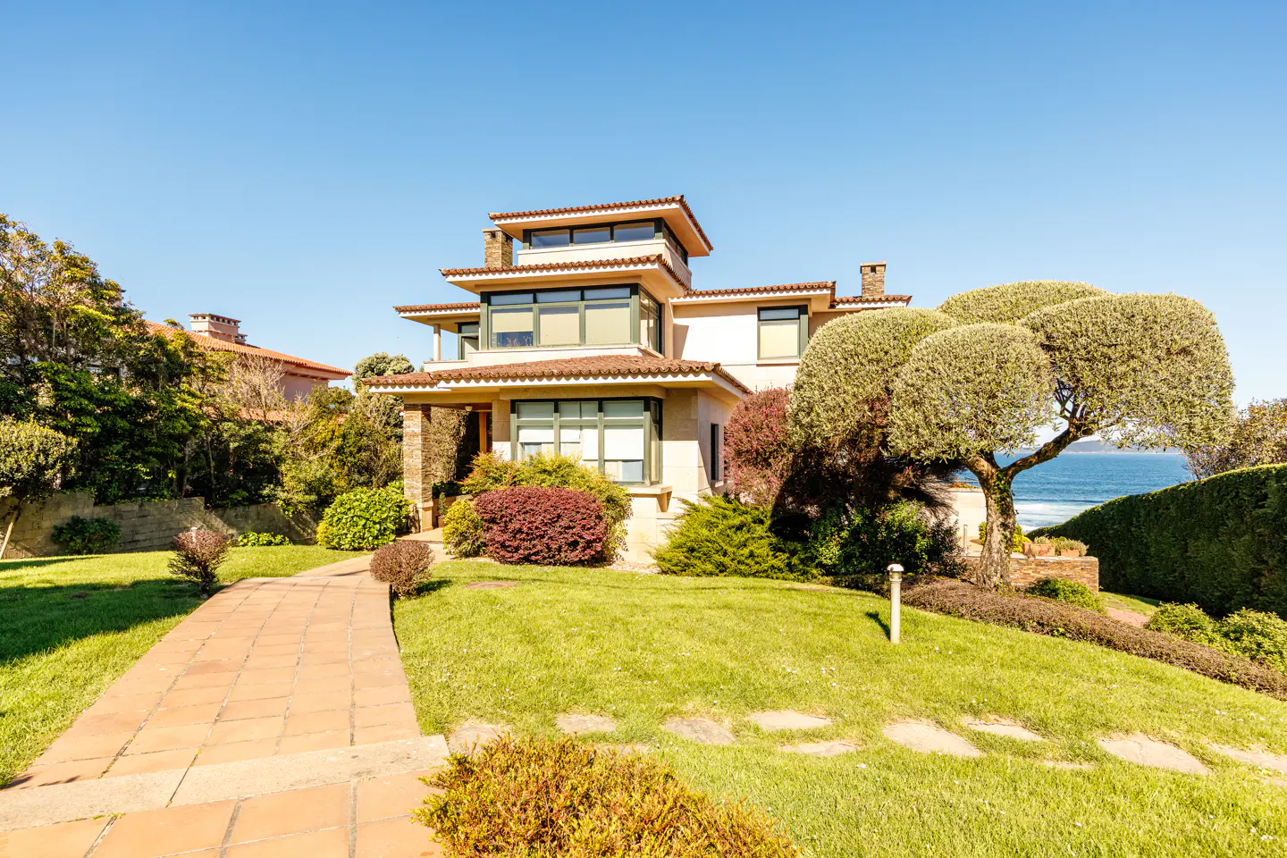 A three-story beige house with green trim, a red tile roof, and a brick walkway on a sunny day. Ocean visible in the background.