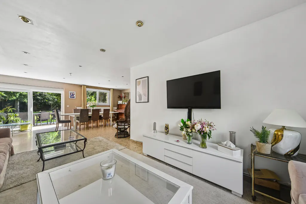 Bright living room with white walls, a large TV, and a white console table with flowers. A dining area is visible in the background.
