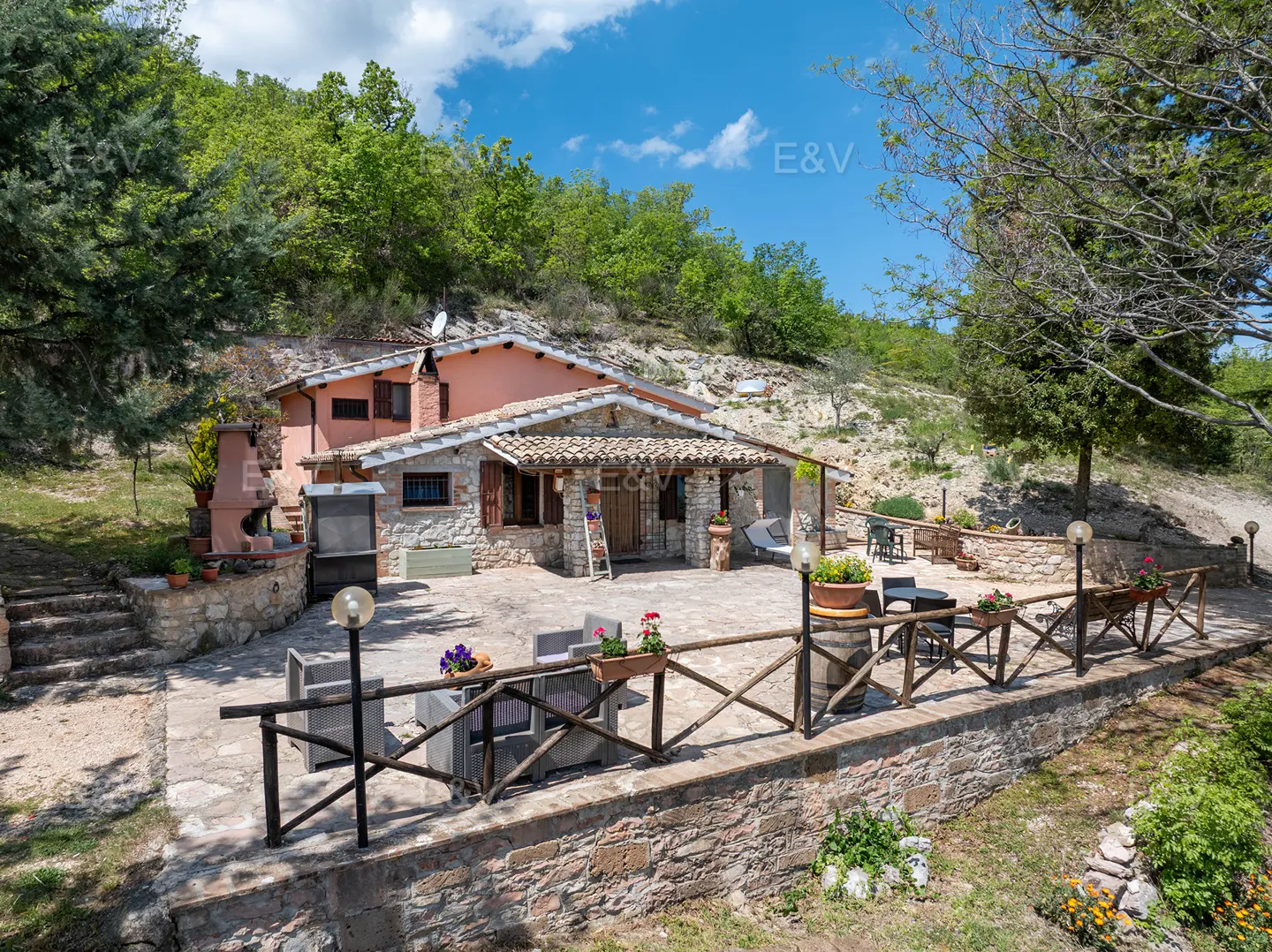 Exterior view of a stone house with a pink section, patio with furniture, and a hillside backdrop.
