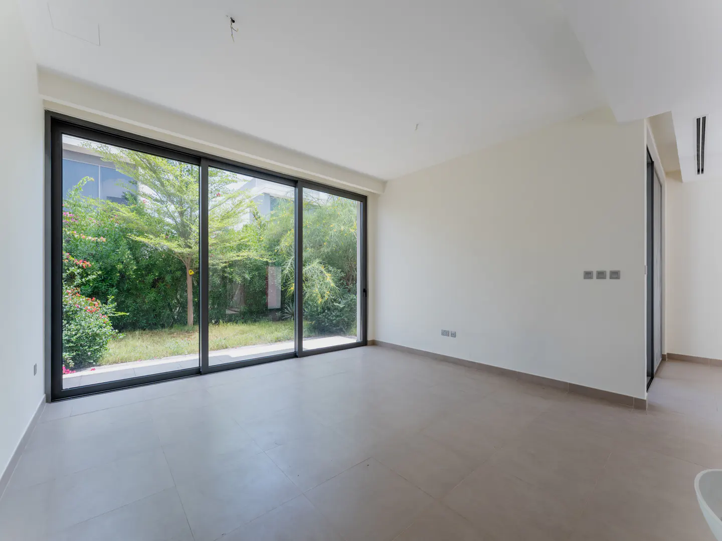 Empty room with beige tile floor, white walls, and large sliding glass doors to a green garden.