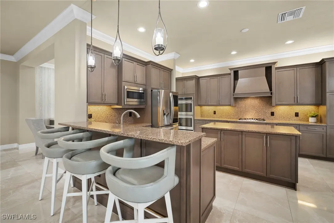A modern kitchen with gray cabinets, granite countertops, and stainless steel appliances. Three pendant lights hang above the island with white stools.