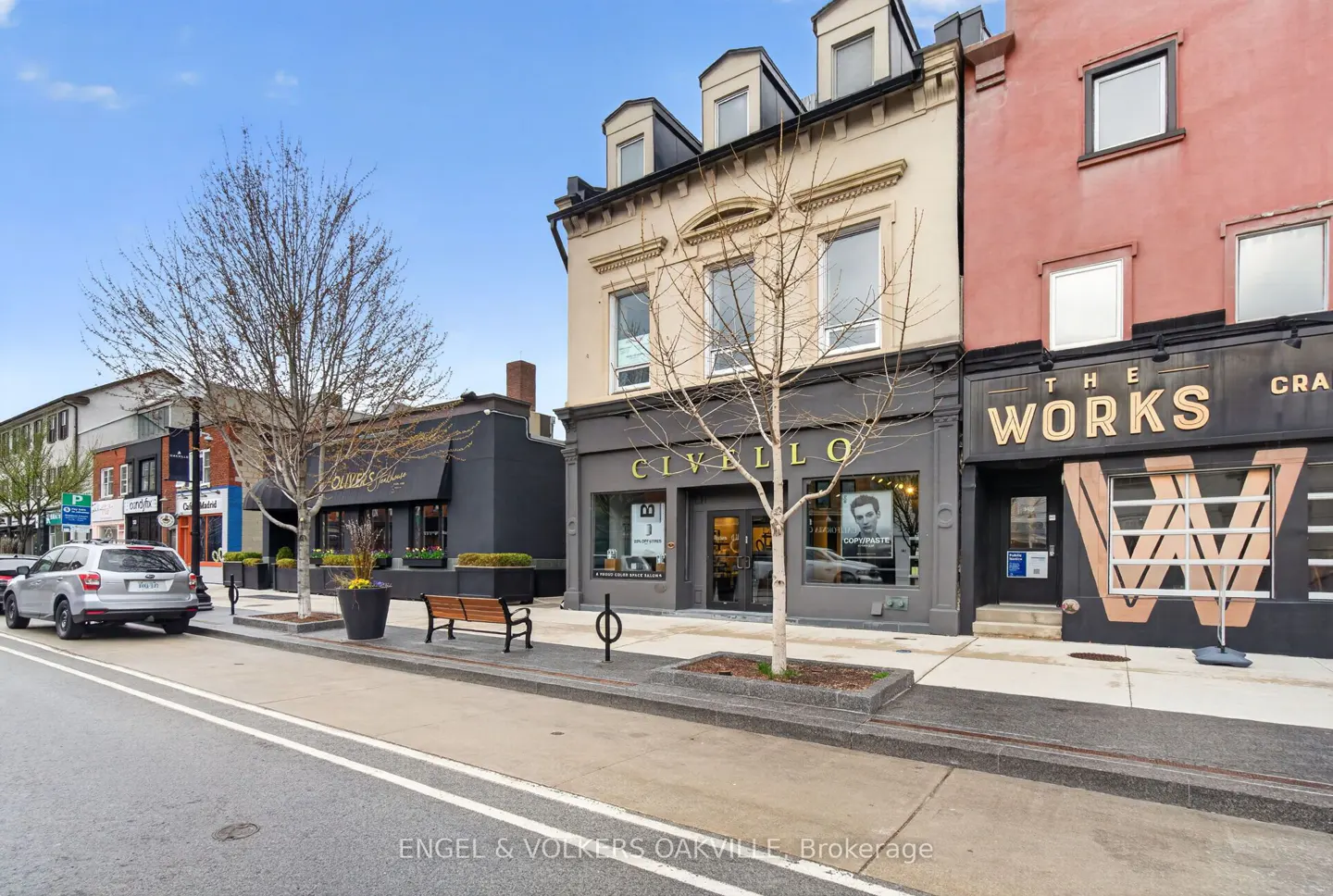 Street view of shops: Civello, The Works, and Oliver's. Bare trees line the sidewalk. A silver car is parked on the street.
