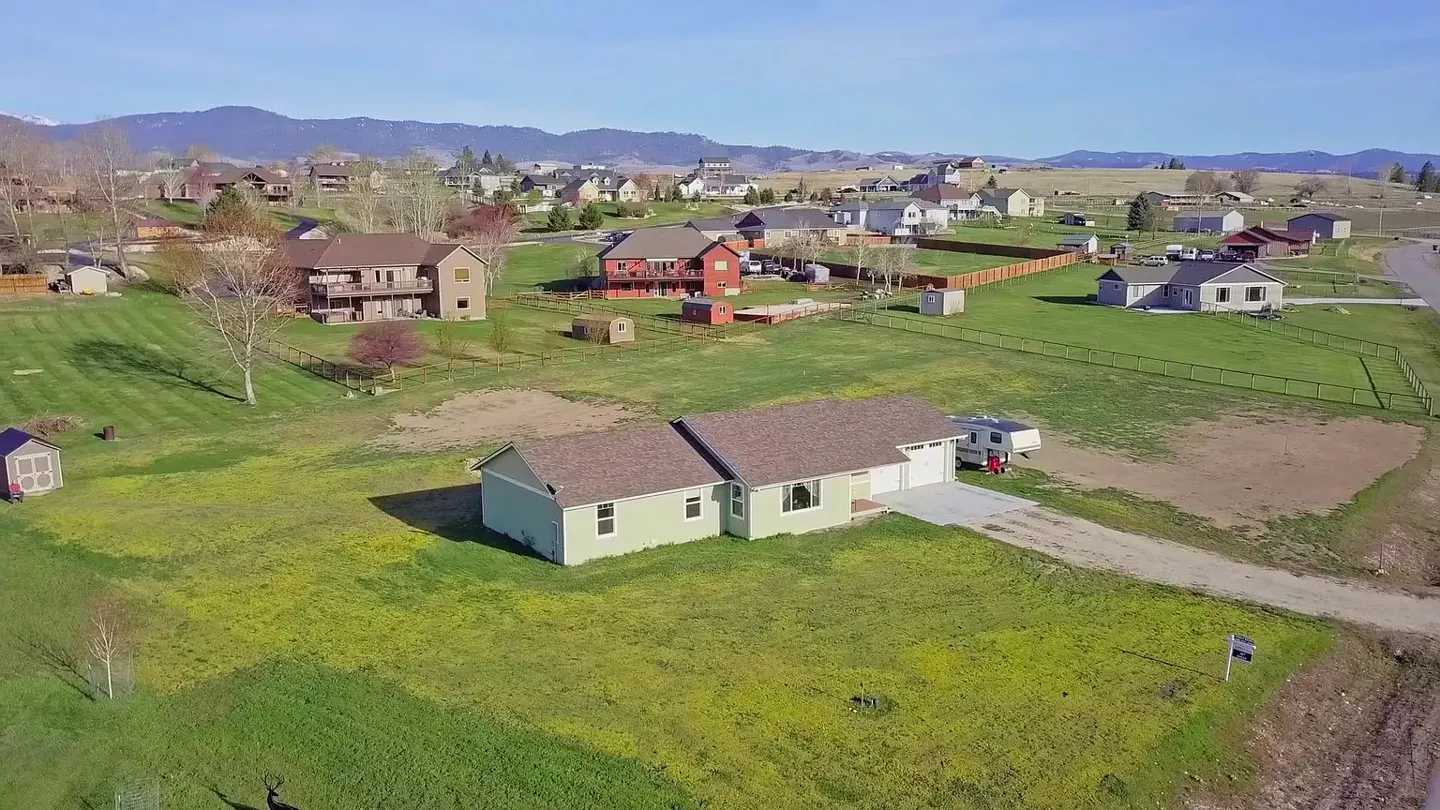 Aerial view of a light green ranch-style house with a brown roof, a camper parked in the driveway, and a large green lawn.