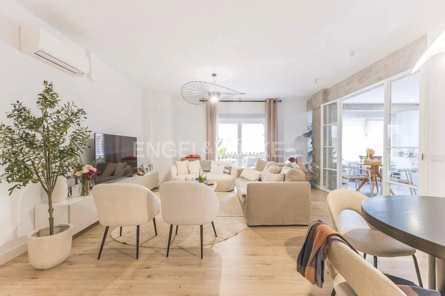 Bright living room with beige sofas, chairs, and wood floors. A large TV sits on a white console table next to a potted plant. Glass doors lead to a dining area.